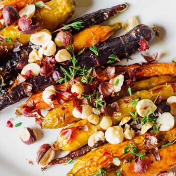 A plate of roasted carrots in various colors, topped with chopped hazelnuts and sprigs of fresh thyme. The dish is presented on a white plate.