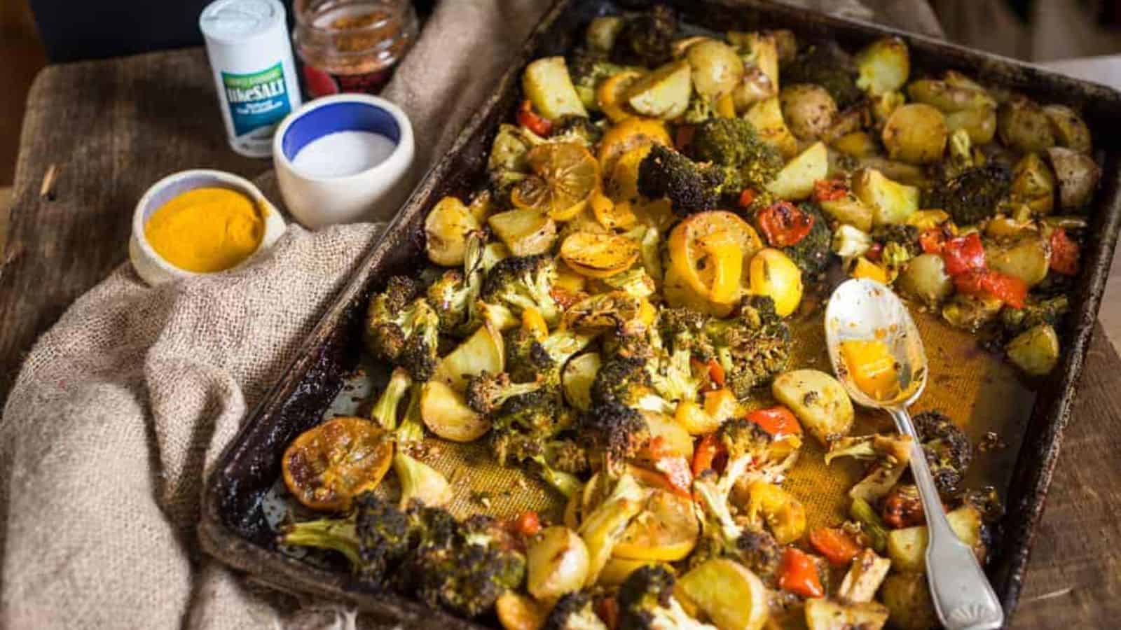 A baking tray filled with roasted mixed vegetables, including potatoes, broccoli, peppers, and sliced lemons, rests on a wooden table. A spoon is on the tray. Nearby are small bowls of spices and a container of sea salt.