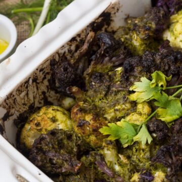 A casserole dish filled with roasted cauliflower topped with green herb sauce. A sprig of parsley is placed on top. A small white cup containing olive oil is on the side, with fresh green herbs scattered on a burlap surface nearby.