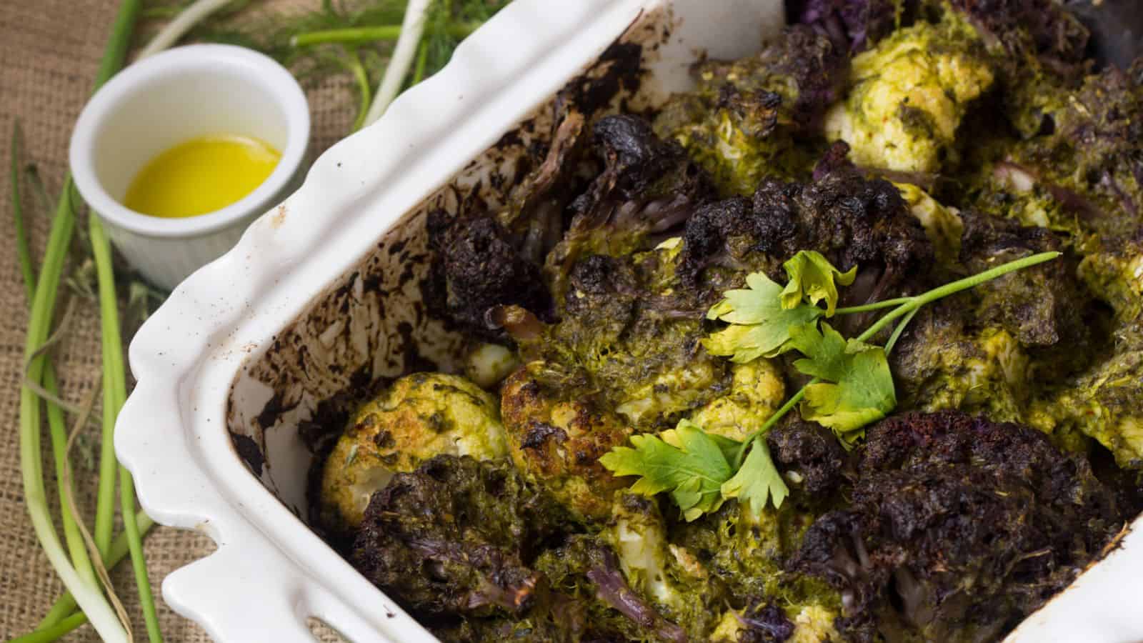 A baked dish with roasted cauliflower florets covered in a green herb sauce is displayed in a white ceramic baking dish. A sprig of fresh parsley garnishes the top. A small bowl of olive oil and a few scallions are placed beside the dish.