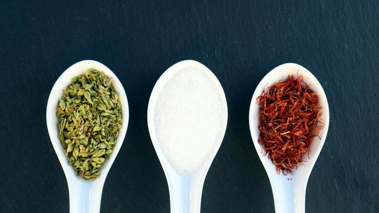 Three white spoons are arranged side by side on a dark surface. The left spoon contains dried green herbs, the middle spoon holds white granular salt, and the right spoon is filled with dried red saffron threads.