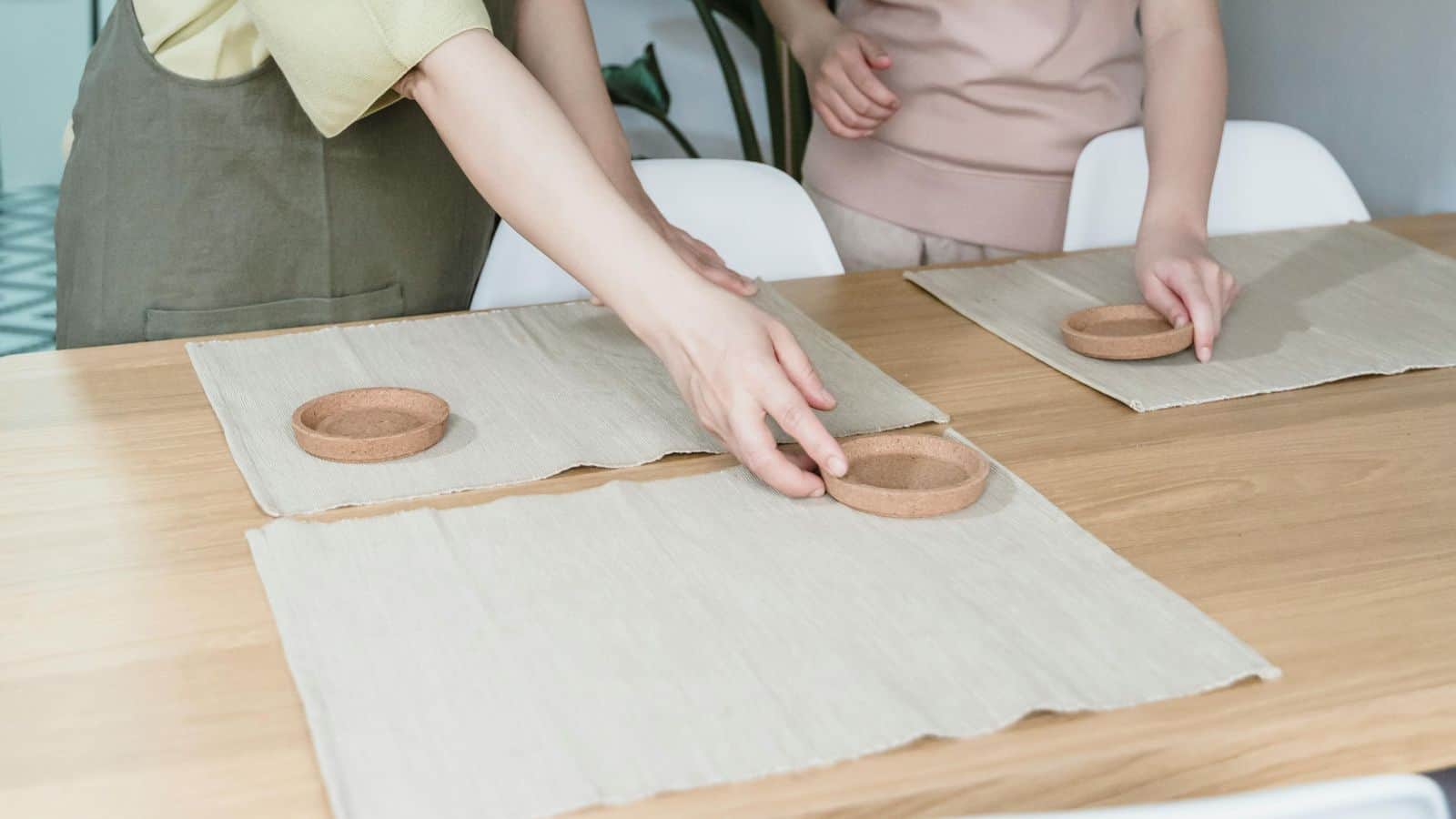 Two people are setting a wooden table with light-colored placemats and round coasters, inspired by holiday meal planning tips. One is reaching over the table while the other places their coaster. Both wear light-colored clothing, creating a serene and stylish ambiance.