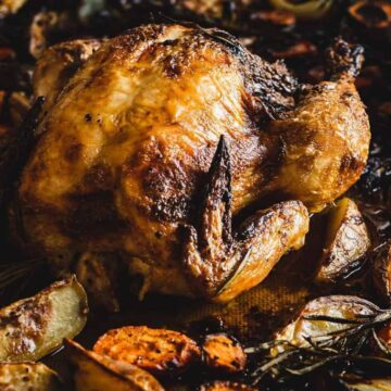 A roasted chicken on a tray surrounded by roasted vegetables, such as onions, carrots, and herbs. Two hands hold the tray at the edges. The chicken is golden-brown and crispy.