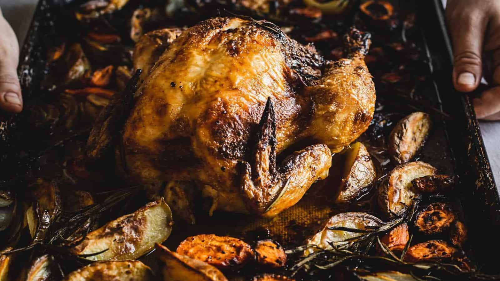 A roasted chicken on a tray surrounded by roasted vegetables, such as onions, carrots, and herbs. Two hands hold the tray at the edges. The chicken is golden-brown and crispy.