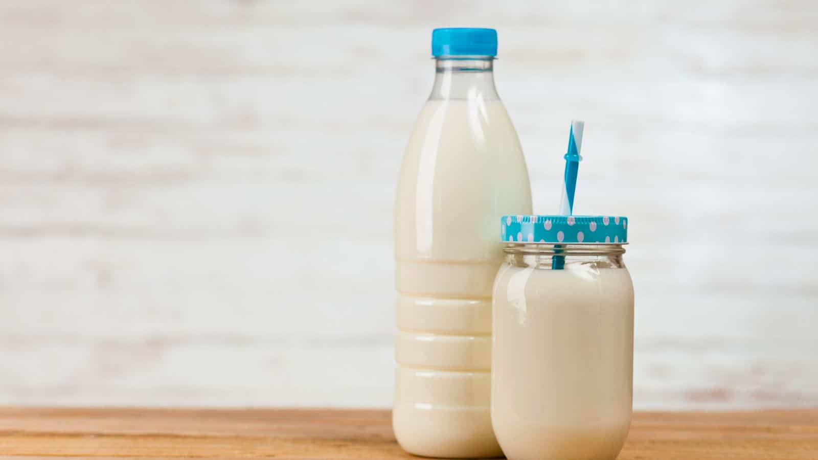 A plastic bottle and a glass jar of milk on a wooden surface. The jar has a blue straw through a polka dot lid. The background is blurred and light-colored, giving a neutral setting.