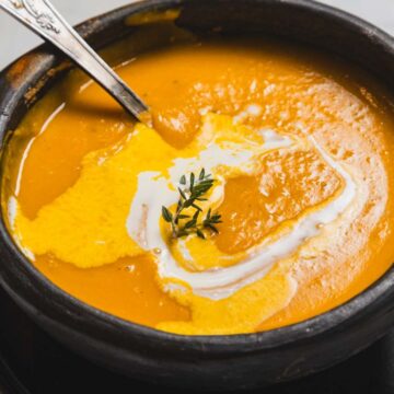 A black bowl filled with creamy orange soup, topped with a swirl of cream and garnished with a sprig of thyme. A silver spoon rests in the bowl. The bowl is placed on a matching black plate on a marble surface.