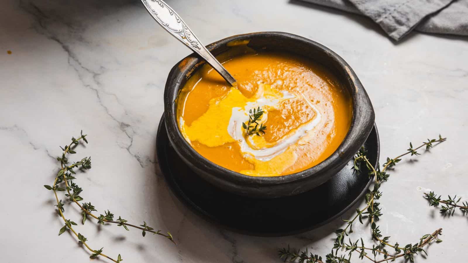 A black bowl with creamy orange soup, garnished with a swirl of cream and a sprig of thyme, sits on a black plate. A silver spoon is placed inside the bowl. Several thyme sprigs are scattered around on a white marble surface.