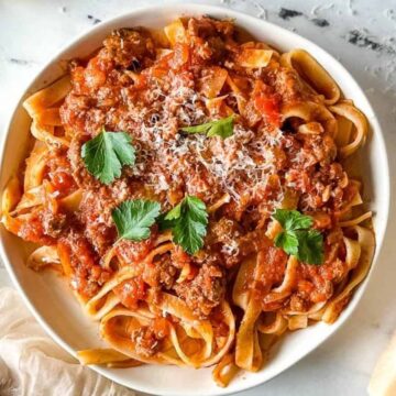 A plate of pasta topped with a tomato and meat sauce, garnished with grated cheese and parsley. A block of cheese, a fork, and a beige napkin are on the marble surface beside the plate.