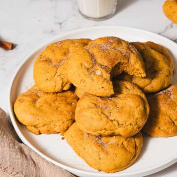 A white plate filled with pumpkin cookies sprinkled with sugar, stacked randomly. One cookie has a bite taken out. A glass of milk and cinnamon sticks are in the background on a marble surface. A beige cloth napkin lies next to the plate.