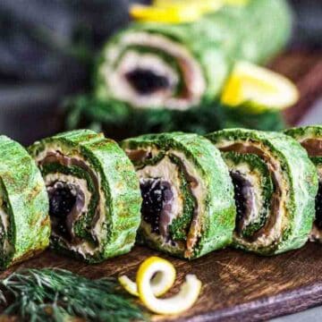 Sliced green roulade on a wooden board, featuring a spiral pattern with layers of filling, possibly including dark and light ingredients. Garnished with fresh herbs and lemon slices in the background.