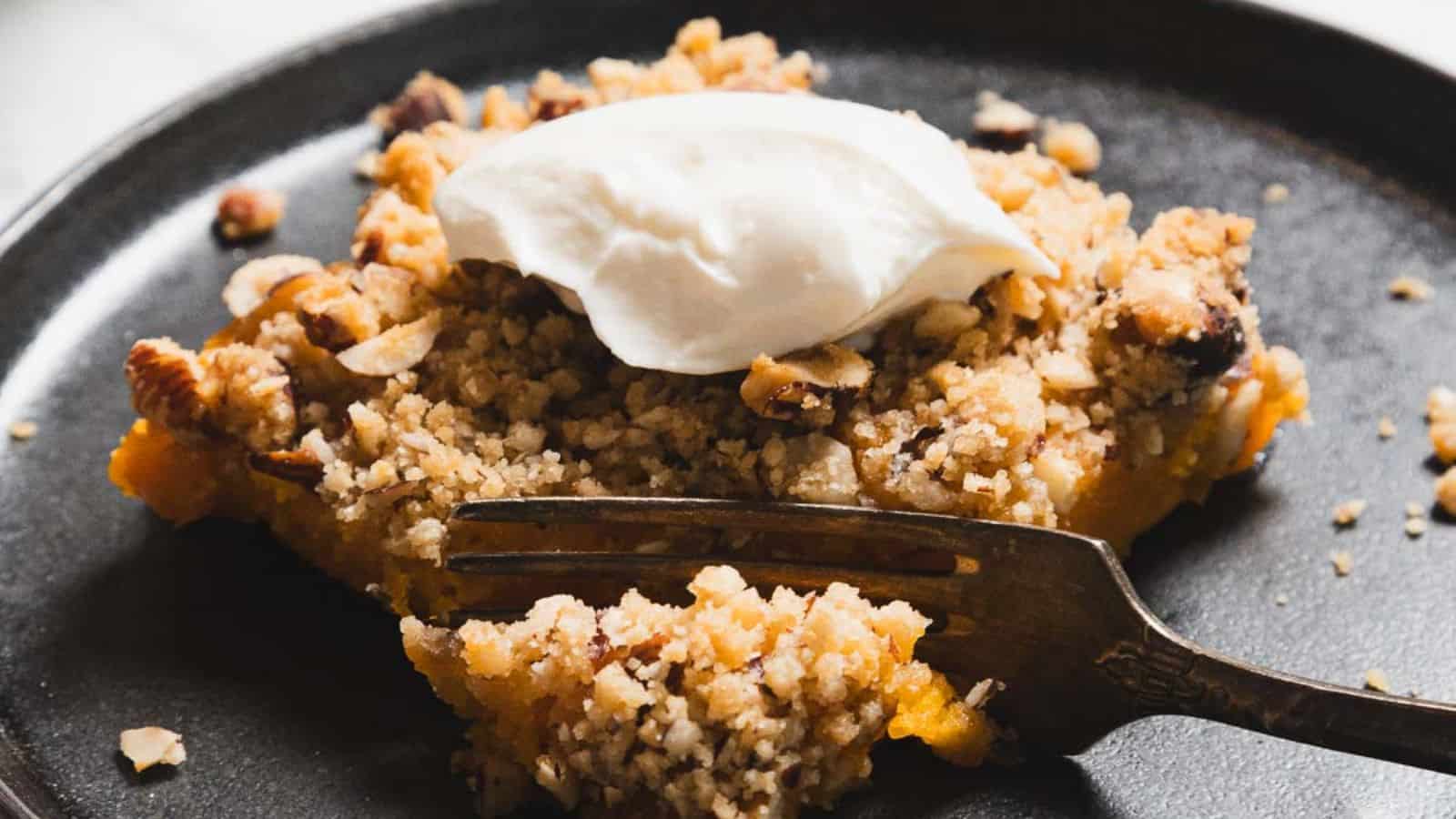 A piece of crumbly dessert with a golden-brown topping is on a dark plate. It has a dollop of whipped cream on top. A fork is partially embedded in the slice, with some crumbs scattered around.