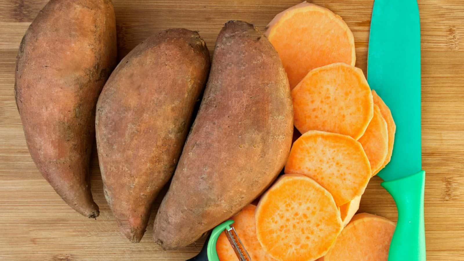 Three whole sweet potatoes and several round slices are placed on a wooden cutting board. A green-handled knife and peeler are also visible next to the sweet potatoes.