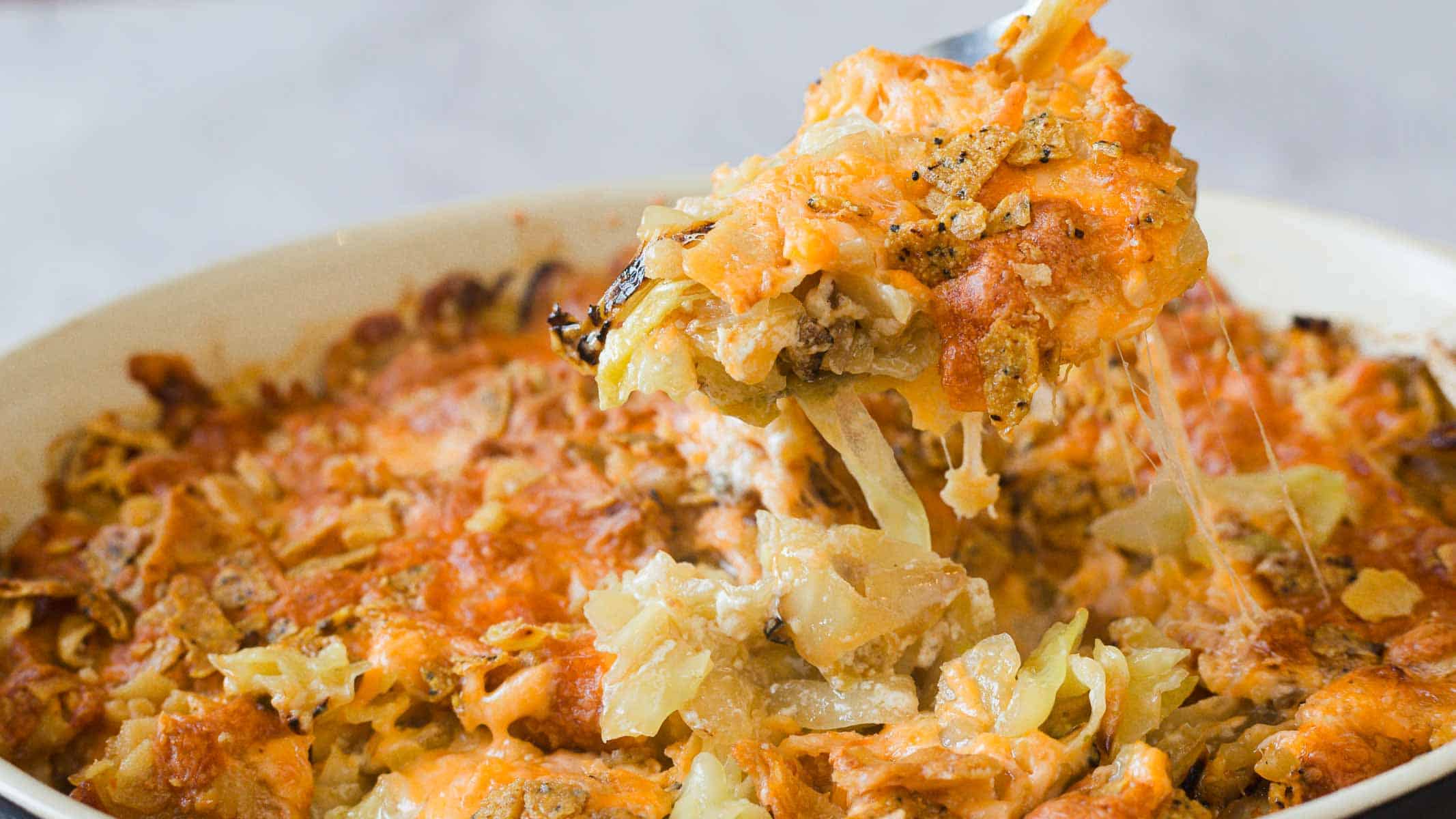 A close-up image of a casserole dish filled with a cheesy cabbage casserole. The top is golden brown and crispy, with a serving spoon lifting a portion, revealing melted cheese and tender cabbage underneath.