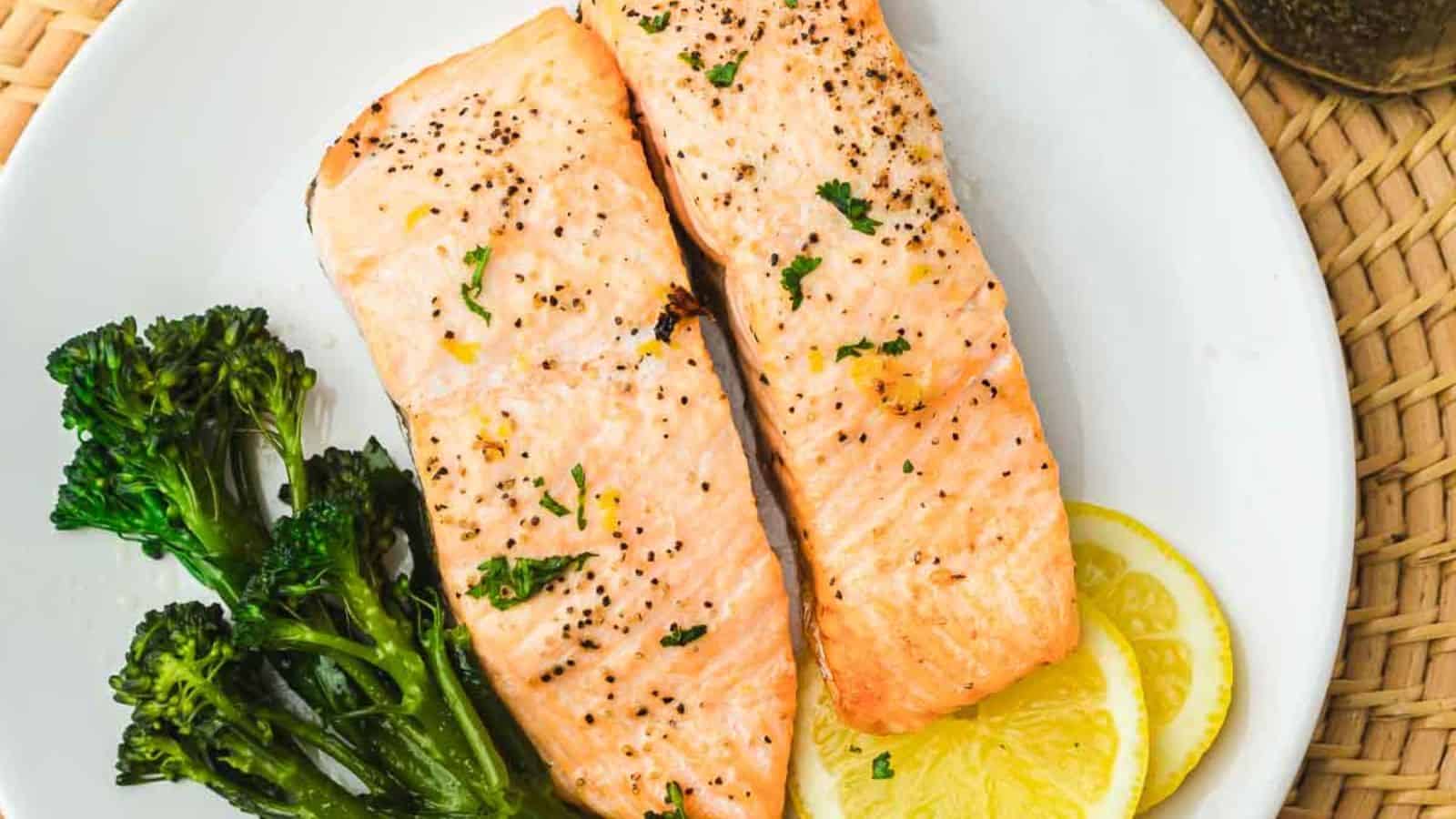 Two pieces of cooked salmon seasoned with black pepper and garnished with herbs are placed on a white plate. The plate also contains steamed broccolini and lemon slices. The background includes a woven placemat.