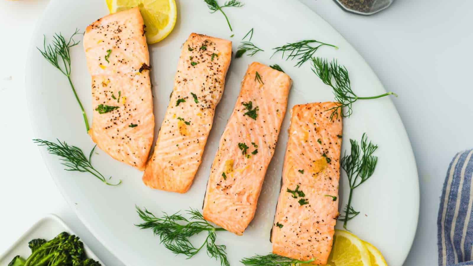 Four grilled salmon fillets garnished with fresh dill and lemon slices are arranged on a white oval platter. The fillets are seasoned with herbs and pepper. The edge of a striped cloth and a serving of broccoli are partially visible.