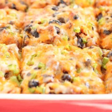 A close-up of a baked casserole in a red dish, featuring melted cheese, black beans, chopped green onions, and diced vegetables. The dish appears freshly cooked and is portioned into squares.