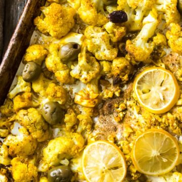 A roasted cauliflower dish on a baking sheet, garnished with lemon slices and olives. A small bowl of green olives and a dish with a sauce are visible in the background on a burlap cloth.