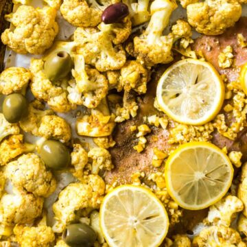 A baking sheet with roasted cauliflower florets, lemon slices, and olives surrounding a piece of salmon. A small bowl of olives and another with sauce are beside the tray. The scene is set on a burlap surface.