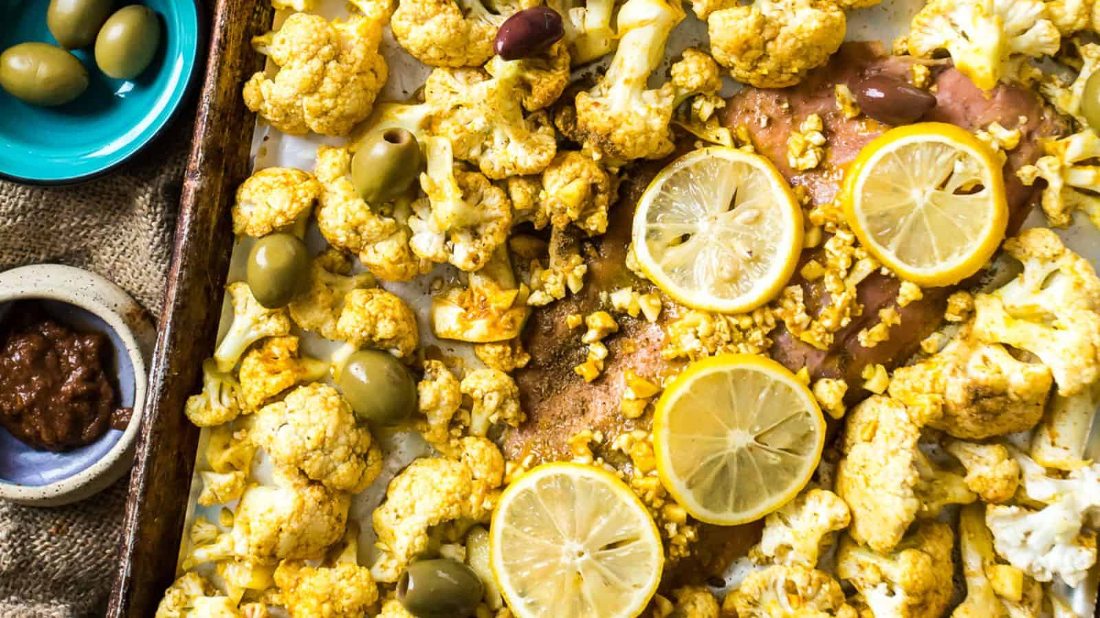 A baking tray displays roasted cauliflower florets, lemon slices, and olives alongside a piece of salmon. A small bowl of olives and another with a dark sauce are placed nearby on a burlap-covered surface.