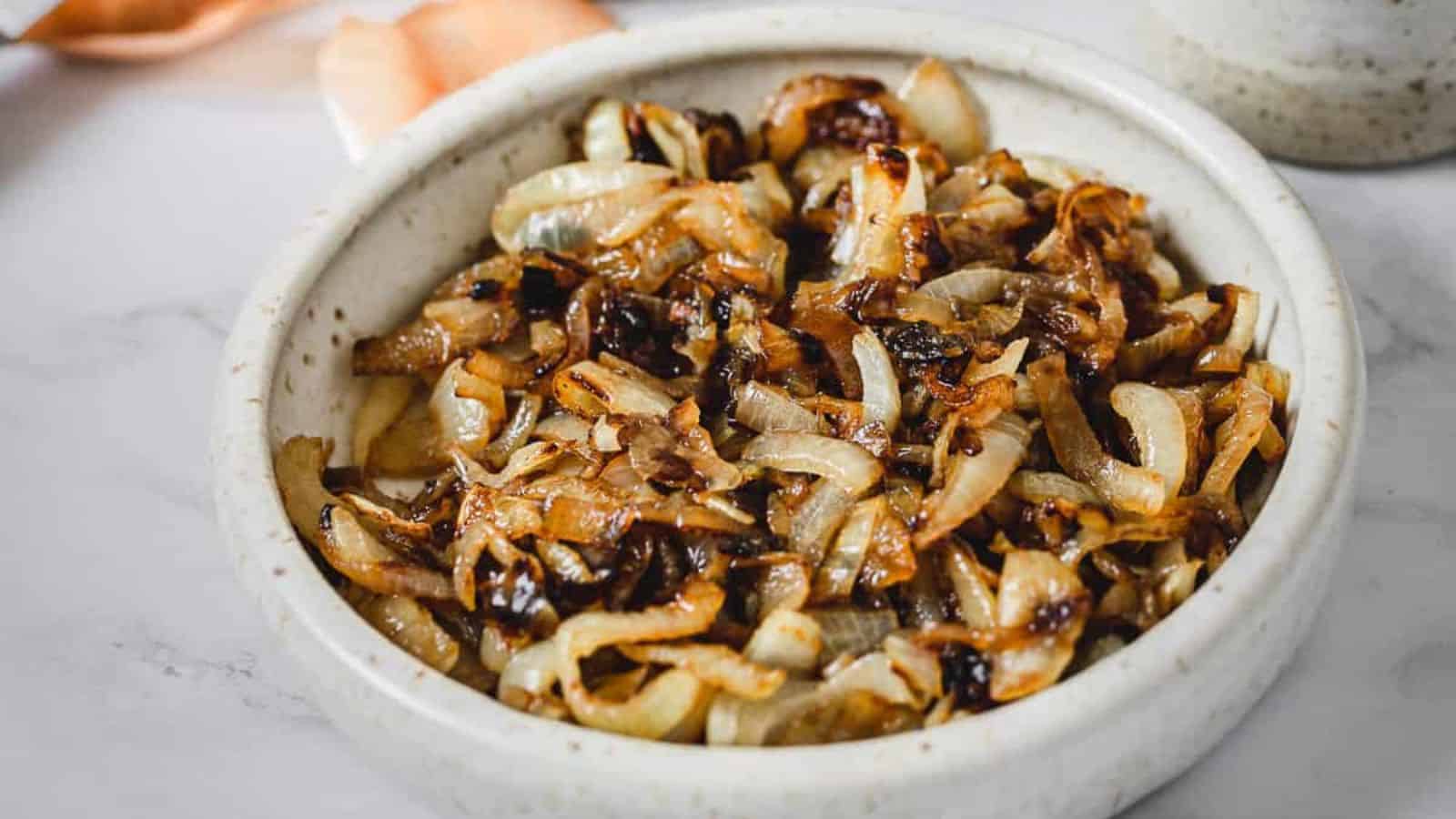 A white ceramic bowl filled with golden brown caramelized onions. The onions are thinly sliced and cooked to a soft texture with a few darker, crispy pieces. The bowl is placed on a marble surface. In the background, a hint of another dish is visible.