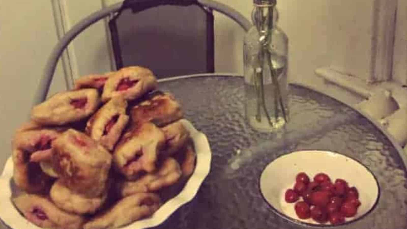 A glass-top table with a plate of pastries stacked on the left, a small bowl of red berries on the right, and a tall bottle with a few green stems in the center.