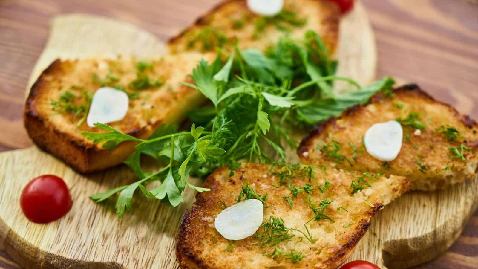 Slices of toasted bread topped with fresh herbs and thin garlic slices are arranged on a wooden board. Cherry tomatoes and a bunch of flat-leaf parsley accompany the toast. The board is placed on a wooden surface.