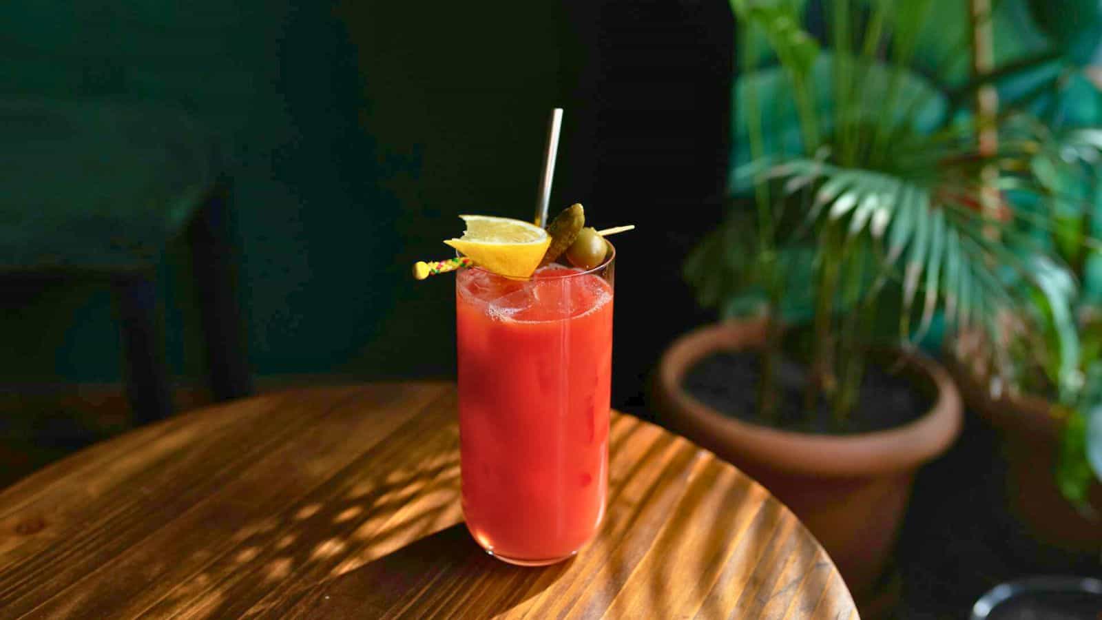 A tall glass of red cocktail with ice, garnished with a lemon wedge and olives on a stick, sits on a wooden table. A straw is inserted in the drink. The background features green foliage and a blurred plant pot.