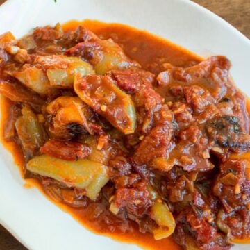A white dish with floral patterns contains a serving of vegetable stew. The stew appears rich and thick, with visible chunks of tomato, onion, and pepper, covered in a red sauce. The dish is set on a wooden surface.