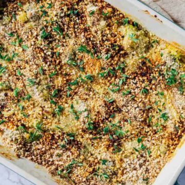 A rectangular baking dish filled with a baked casserole topped with breadcrumbs and fresh herbs. The surface is golden brown and garnished with green leaf sprigs. The dish rests on a marble surface beside a light-colored cloth.
