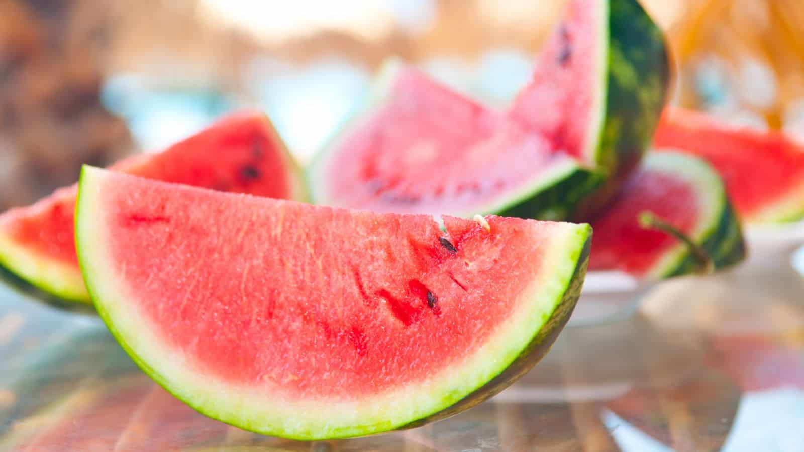 Close-up of several sliced watermelon pieces on a glass surface. The slices display bright red fruit with black seeds and green rind, placed in a way that highlights their juicy texture. The background is softly blurred.