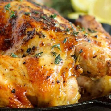 A roasted chicken in a cast iron skillet, garnished with herbs. The chicken has a golden-brown, crispy skin. Slices of lemon and herbs are visible in the background.