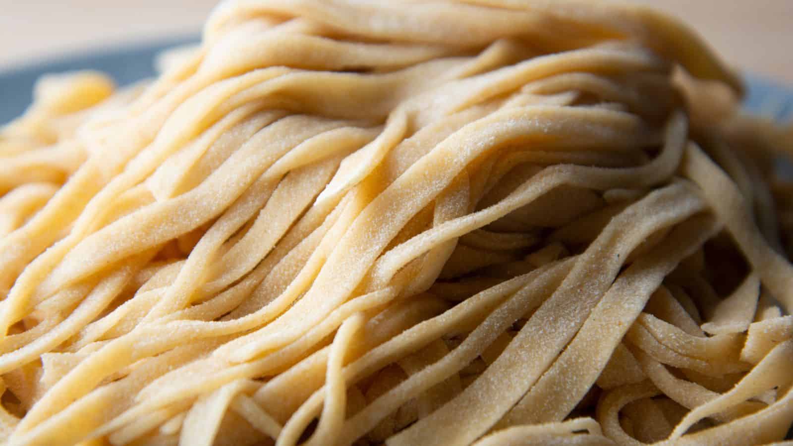 A close-up of a pile of uncooked, fiber-rich fettuccine pasta. The noodles are slightly dusted with flour, enhancing their textured appearance. The image focuses on the golden color and ribbon-like shape of this nutritious pasta.
