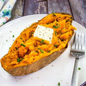 A baked sweet potato, part of this week's delicious menu, is halved on a plate, crowned with a pat of butter and sprinkled with chopped chives and black pepper. A fork rests on the plate beside this delightful dish, while a floral napkin adds charm in the background.