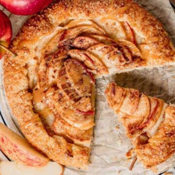 A rustic apple galette with a slice removed is displayed on parchment paper. Perfect for Thanksgiving, the crust is golden brown, topped with apple slices and a drizzle of caramel. Whole red apples and cinnamon sticks are nearby on a wire cooling rack.