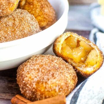 A bowl of cinnamon sugar-coated apple fritters rests on a wooden surface. One fritter is cut open to reveal apple chunks, making it perfect for daily baking enthusiasts. Nearby, cinnamon sticks and a striped cloth add charm, while butter packages peek from the background, hinting at easy recipes to try.