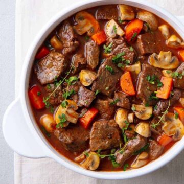 A white bowl filled with beef stew, containing chunks of beef, sliced carrots, mushrooms, and garnished with chopped herbs. The stew is served on a cloth alongside a metallic spoon.