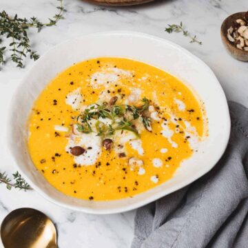 A white bowl filled with creamy butternut squash soup with apples is topped with a drizzle of cream, black pepper, and fresh herbs. Small pieces of nuts are sprinkled on top. A gray cloth and a gold spoon rest beside the bowl on a marble surface.