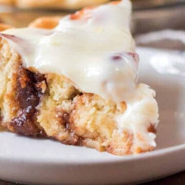 A slice of cinnamon roll cake with cream cheese frosting on a white plate. The cake appears soft and moist, with visible swirls of cinnamon.