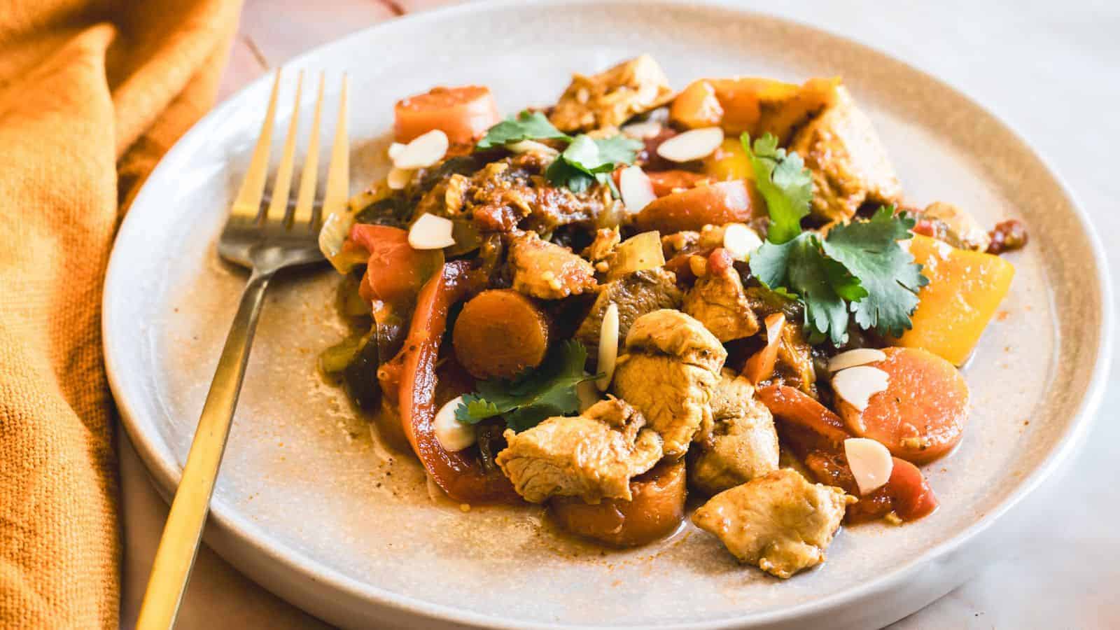 A plate of chicken stir-fry with sliced carrots, bell peppers, and mushrooms, garnished with cilantro and almond slices. A fork rests on the plate, and a yellow cloth is visible in the background.