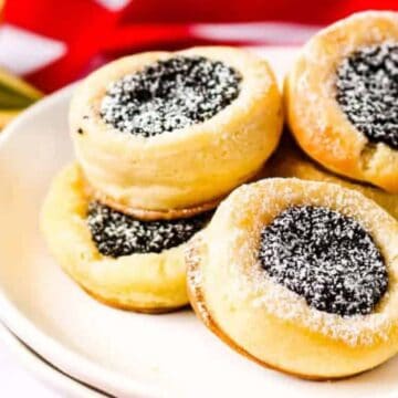 A plate of round pastries filled with a dark filling, sprinkled with powdered sugar. The pastries are stacked on a white plate, with a red and white background and a gold ribbon visible to the side.