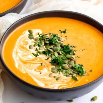 A bowl of creamy pumpkin soup, perfect for Thanksgiving recipes, is garnished with a swirl of cream, fresh parsley, and pumpkin seeds. The bowl rests on a white surface with a light fabric draped nearby. Another bowl of this delightful dish sits partially visible in the background.