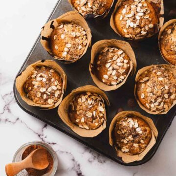 A tray of pumpkin spice muffins, topped with oats and cinnamon, is displayed on a marble surface. A small wooden spoon and a jar of cinnamon sit beside the tray, adding to the cozy autumn vibes. Each muffin is wrapped in brown parchment paper.