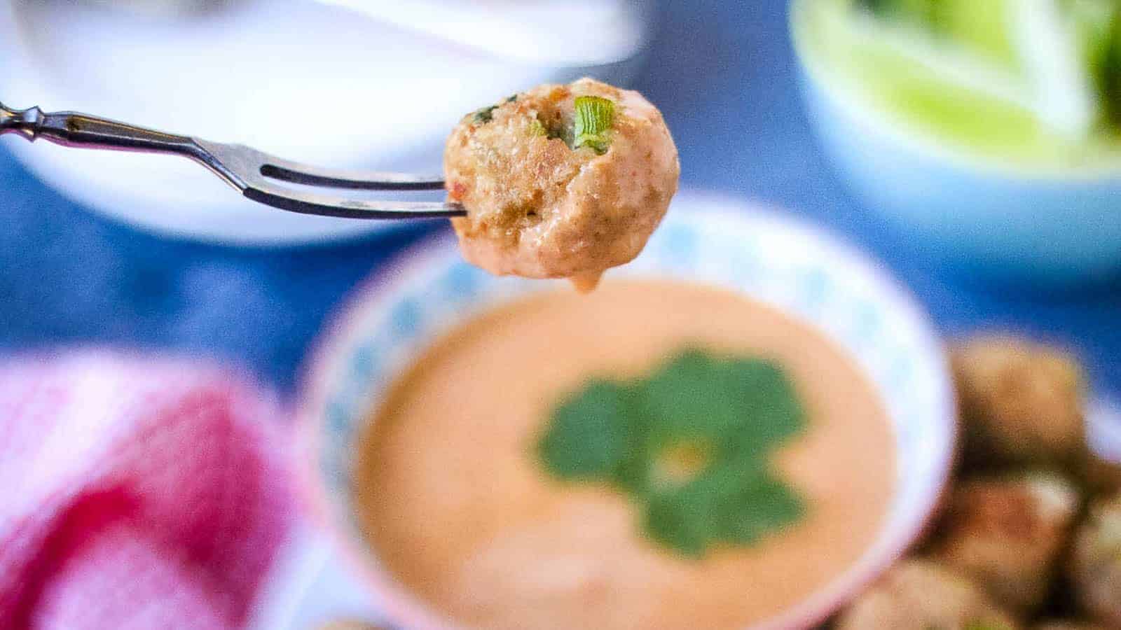 A meatball is held on a fork above a bowl of creamy orange sauce topped with a fresh cilantro leaf, capturing the essence of food photography. The blurred background includes a green salad and a red-checked napkin, creating an auto draft vision for any aspiring SEO-centric culinary blog.