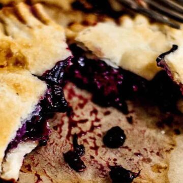 Close-up of a baked hand pie with a flaky golden crust, filled with a juicy dark berry filling. Partially cut open to reveal the rich purple interior, it's placed on brown parchment paper with a fork nearby. Easy pies like this can make every day feel special.
