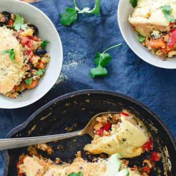 A skillet with sliced cornbread casserole packed with vegetables and beans sits on a wooden table, embodying Thanksgiving dishes that please everyone. Nearby, two bowls filled with the savory casserole rest on a blue cloth, garnished with fresh cilantro.