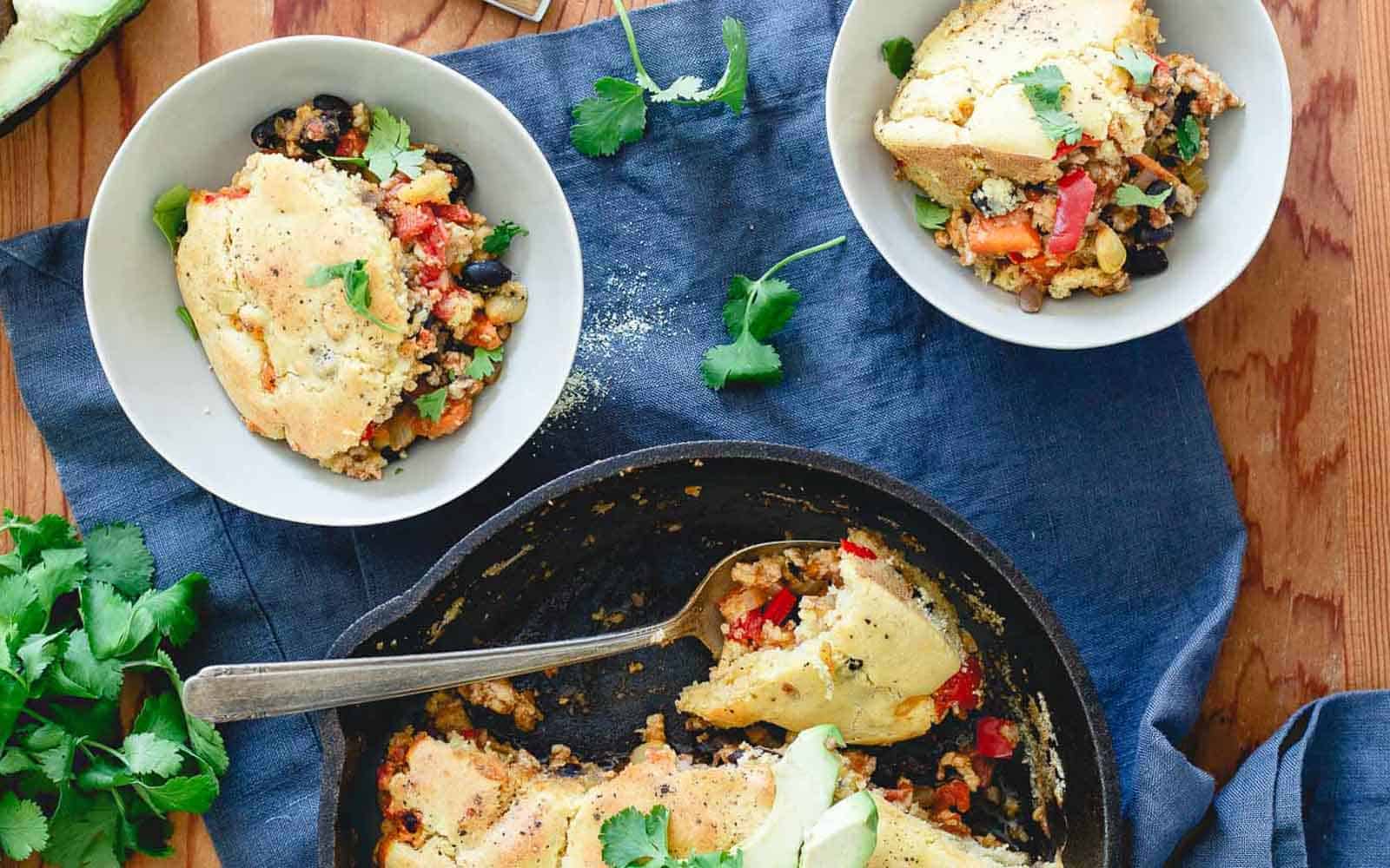 A skillet holds a slice of savory pie, served in two white bowls. This easy recipe includes vegetables and is topped with herbs. Fresh cilantro is scattered around, and a blue cloth is underneath the skillet and bowls—perfect for those who enjoy making homemade desserts like pies.