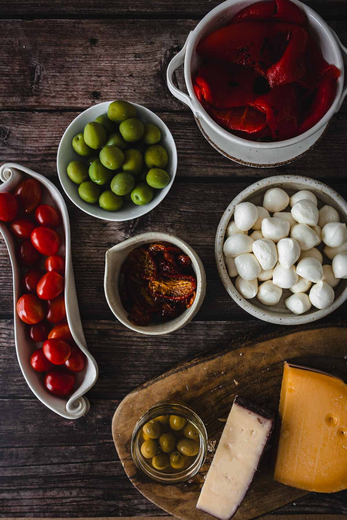 A wooden table displays small bowls of ingredients including green olives, roasted red peppers, mozzarella balls, cherry tomatoes, and sun-dried tomatoes. A wooden board holds cheese wedges and a small bowl of olives.