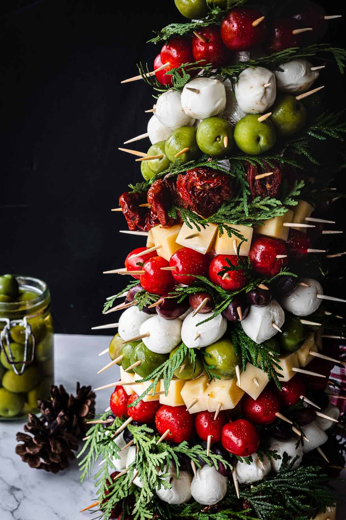 A festive tower of skewered appetizers is shaped like a Christmas tree, featuring tomatoes, mozzarella balls, olives, cheese cubes, and greens. A jar of olives and a pine cone are on the marble surface beside it, set against a dark background.