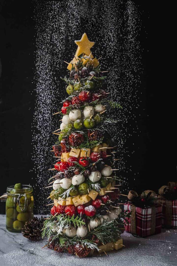 A festive food display featuring a Christmas tree made of skewered cheese, olives, and tomatoes. It is topped with a star-shaped cheese and garnished with herbs. Snow-like powder is sprinkled over it. Pinecones and a jar are nearby on a marble surface.