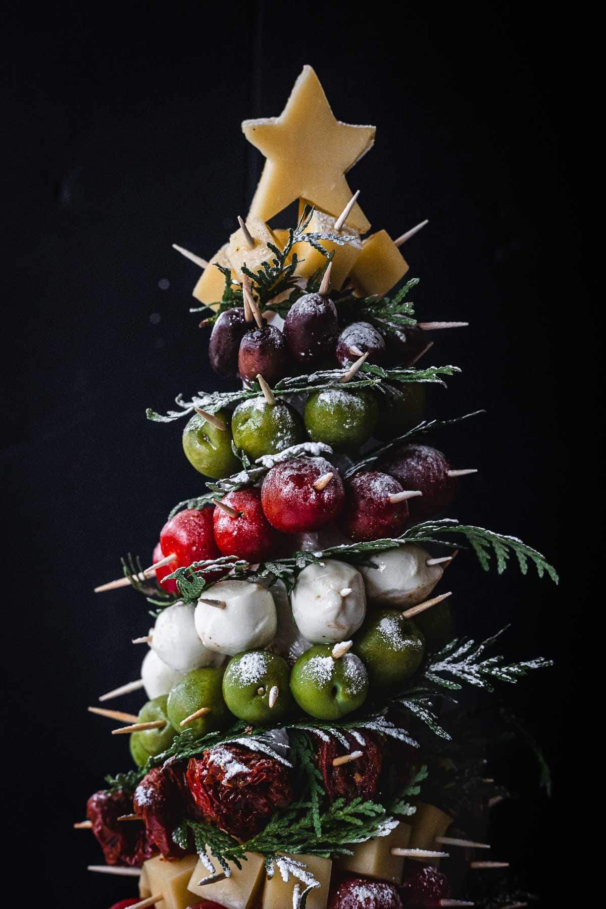 A Christmas tree-shaped food arrangement made with skewers of cheese cubes, olives, cherry tomatoes, mozzarella balls, and herbs, topped with a star-shaped piece of cheese.